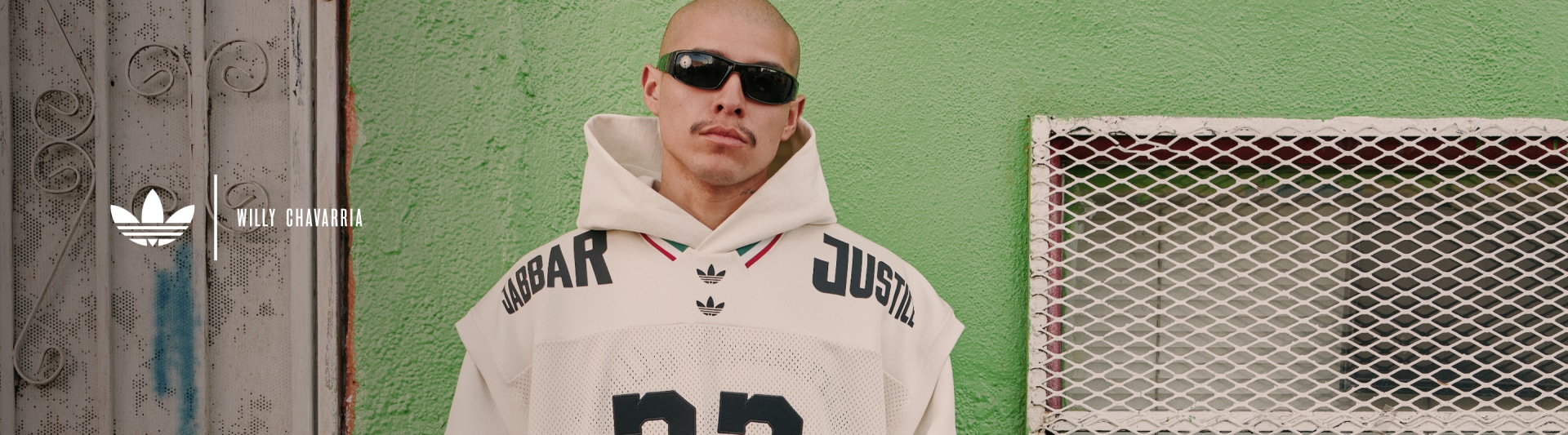 A bald man wearing sunglasses and a white jersey with the word "Adidas" printed on it stands in front of a green wall.