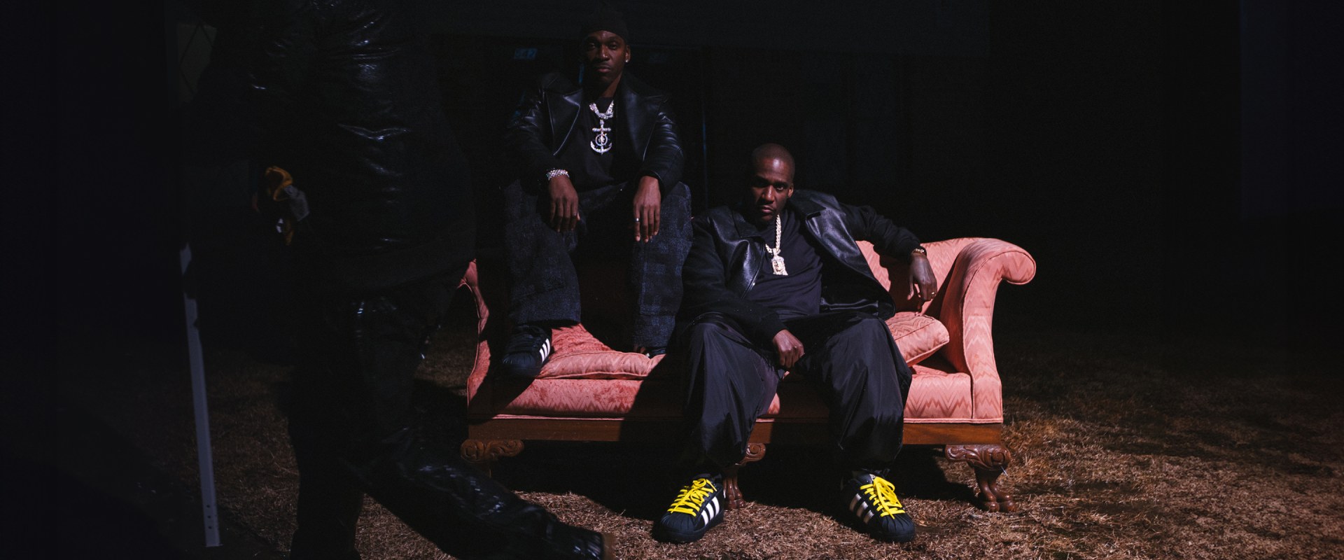 The image shows two individuals sitting on a pink couch in a dark, outdoor setting, with a third person standing behind them.
