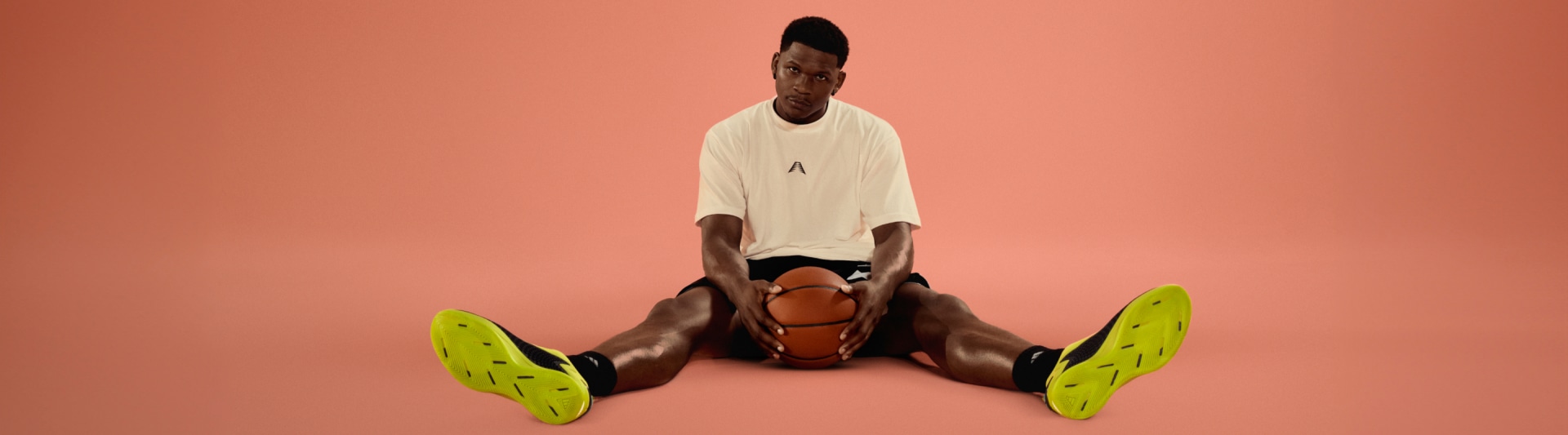 Anthony Edwards wearing a white t-shirt and sitting on the floor, holding a basketball, with bright green sneakers on his feet against a coral-colored background.