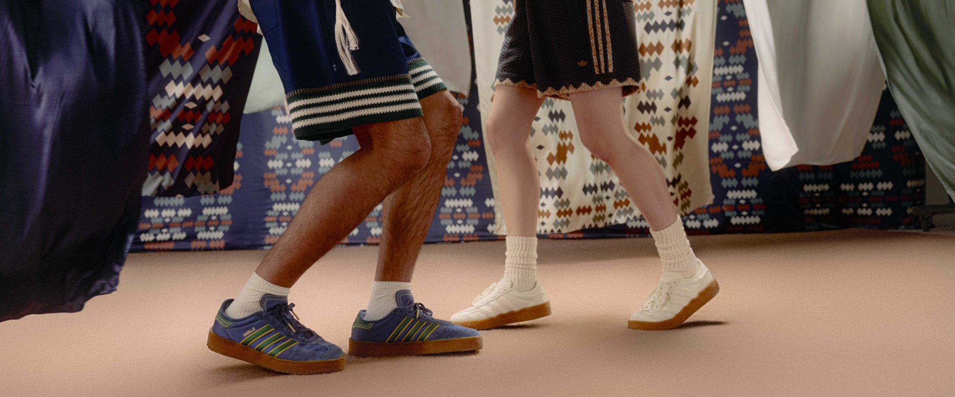 Two people wearing blue and white adidas Edison Chen Silk Gazelle sneakers with gum soles stand on a beige floor. The background features patterned fabric.