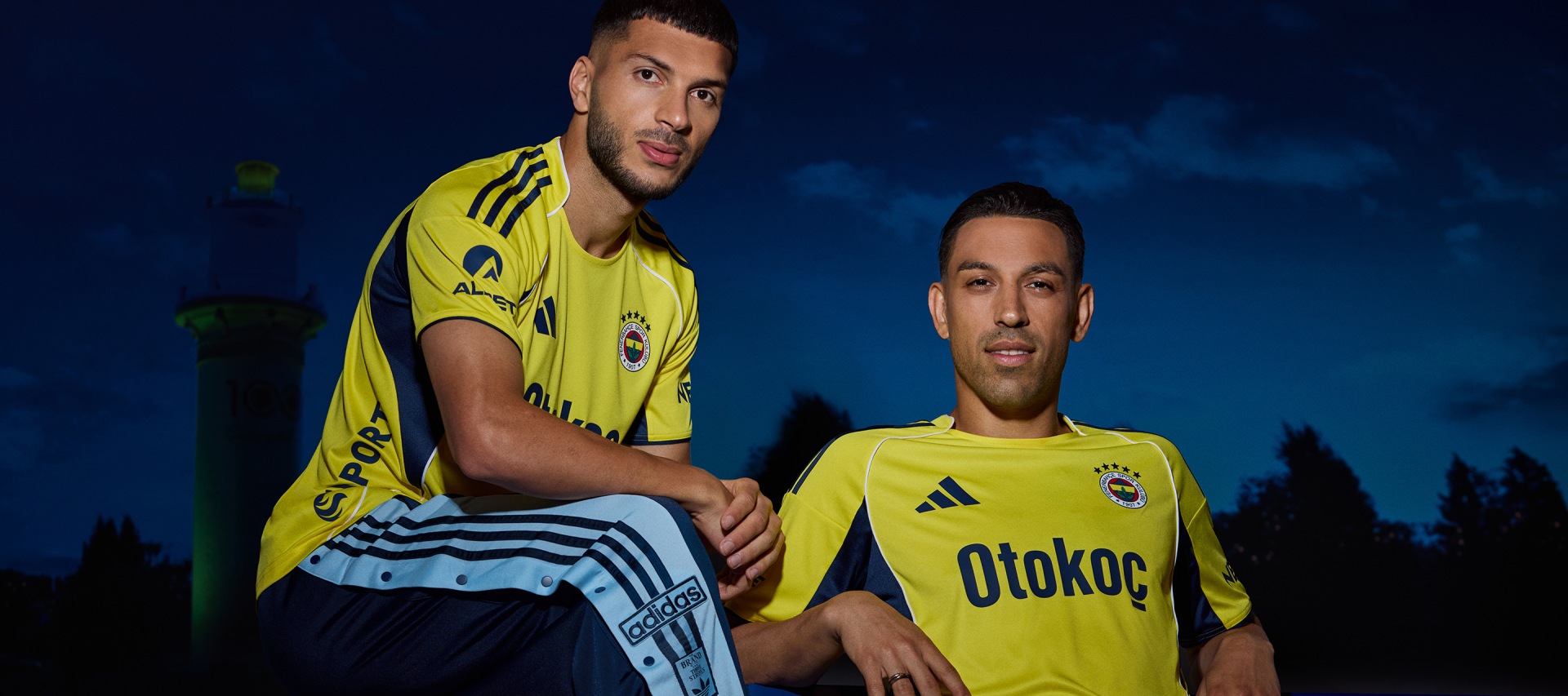 Two men wearing yellow soccer jerseys with the Otokoc logo stand in a night setting with a dark blue sky in the background.