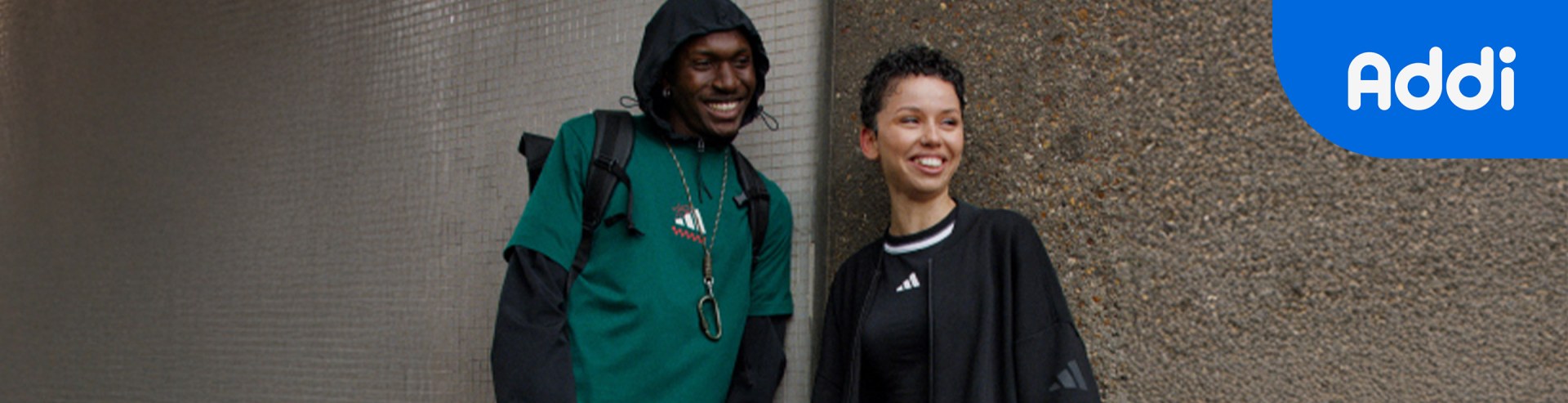 Two individuals, one wearing a green hooded sweatshirt and the other wearing a black jacket, are standing in front of a textured wall.