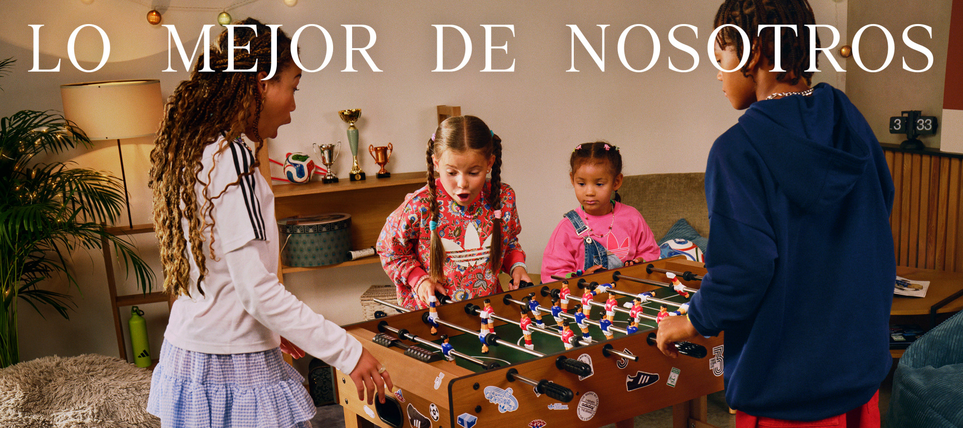The image shows a group of young girls playing a foosball table in what appears to be a home or community setting, with a sign in Spanish that reads "Lo mejor de nosotros" (The best of us) visible in the background.