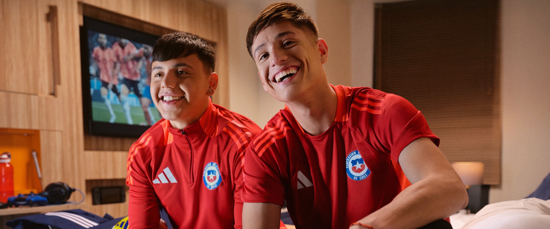 Two young men wearing red soccer jerseys are smiling and posing together in what appears to be a living room or home setting, with a television screen visible in the background.