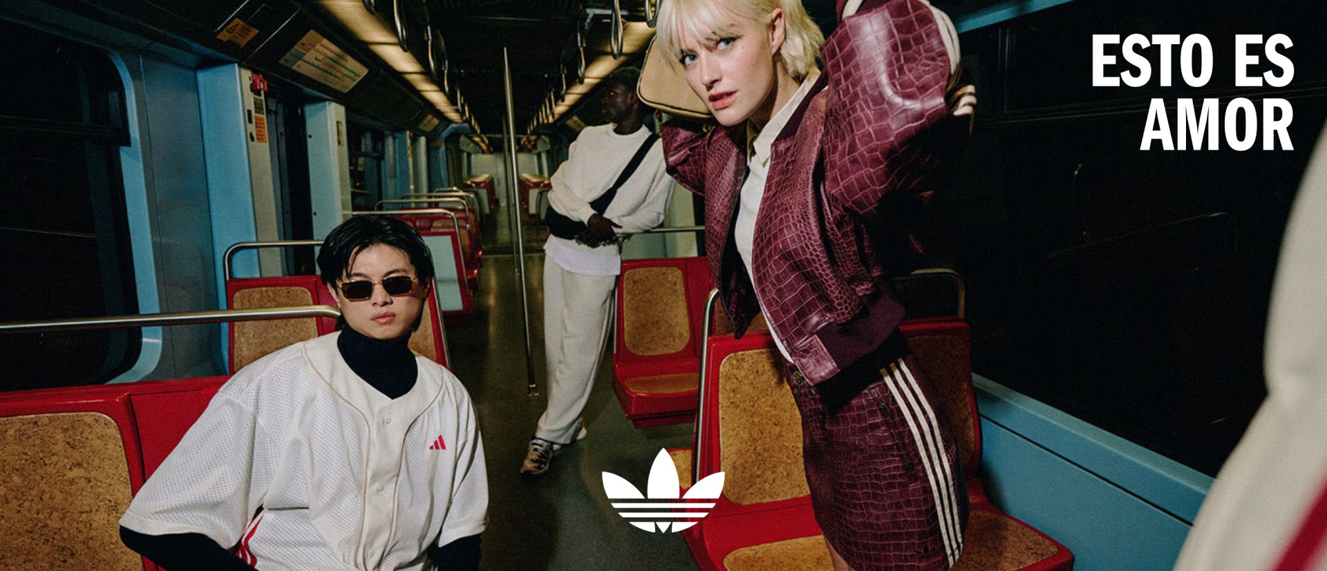 Two individuals, one wearing a white outfit and the other a burgundy outfit, stand in what appears to be a public transportation setting, with colorful seats and a sign in the background.