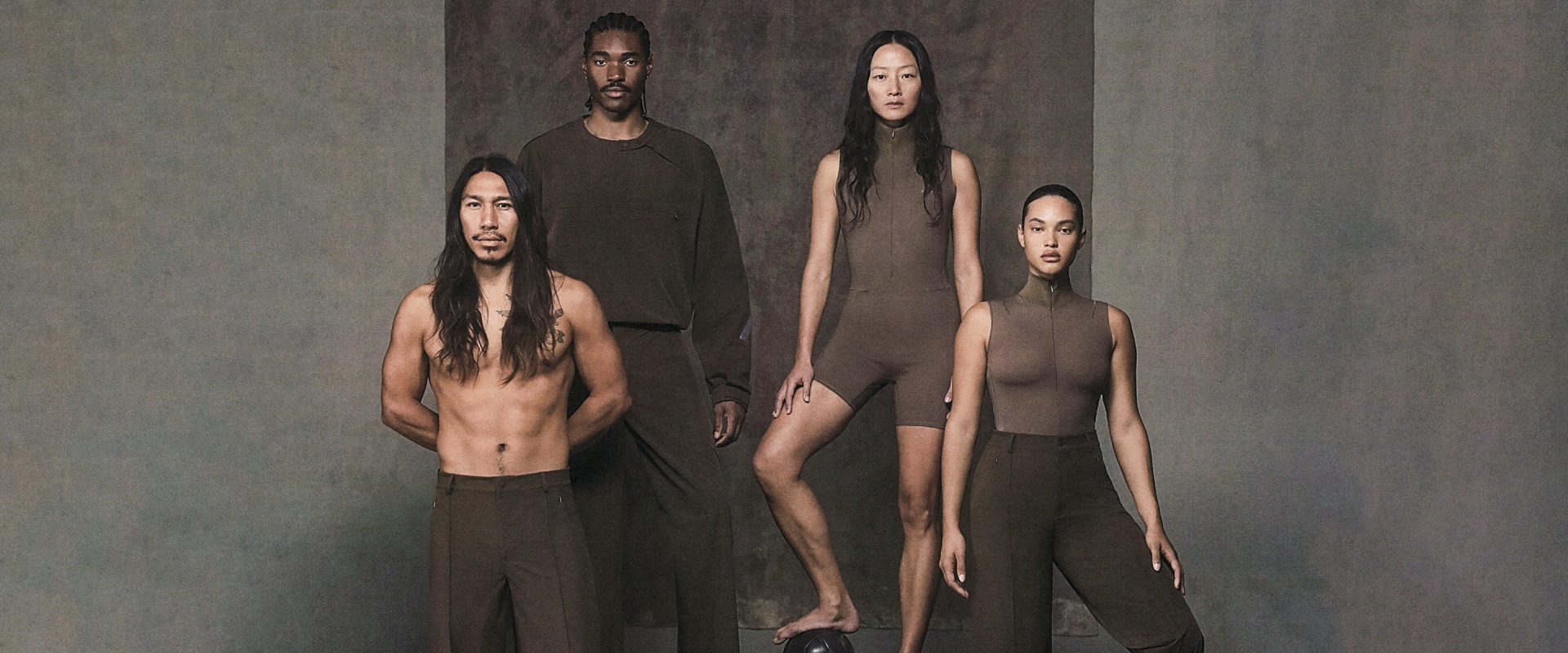 The image features four individuals, two men and two women, standing against a plain, dark background. The individuals are wearing minimal clothing, with the men in dark tops and the women in nude-colored garments. Their poses and expressions convey a sense of contemplation or introspection.