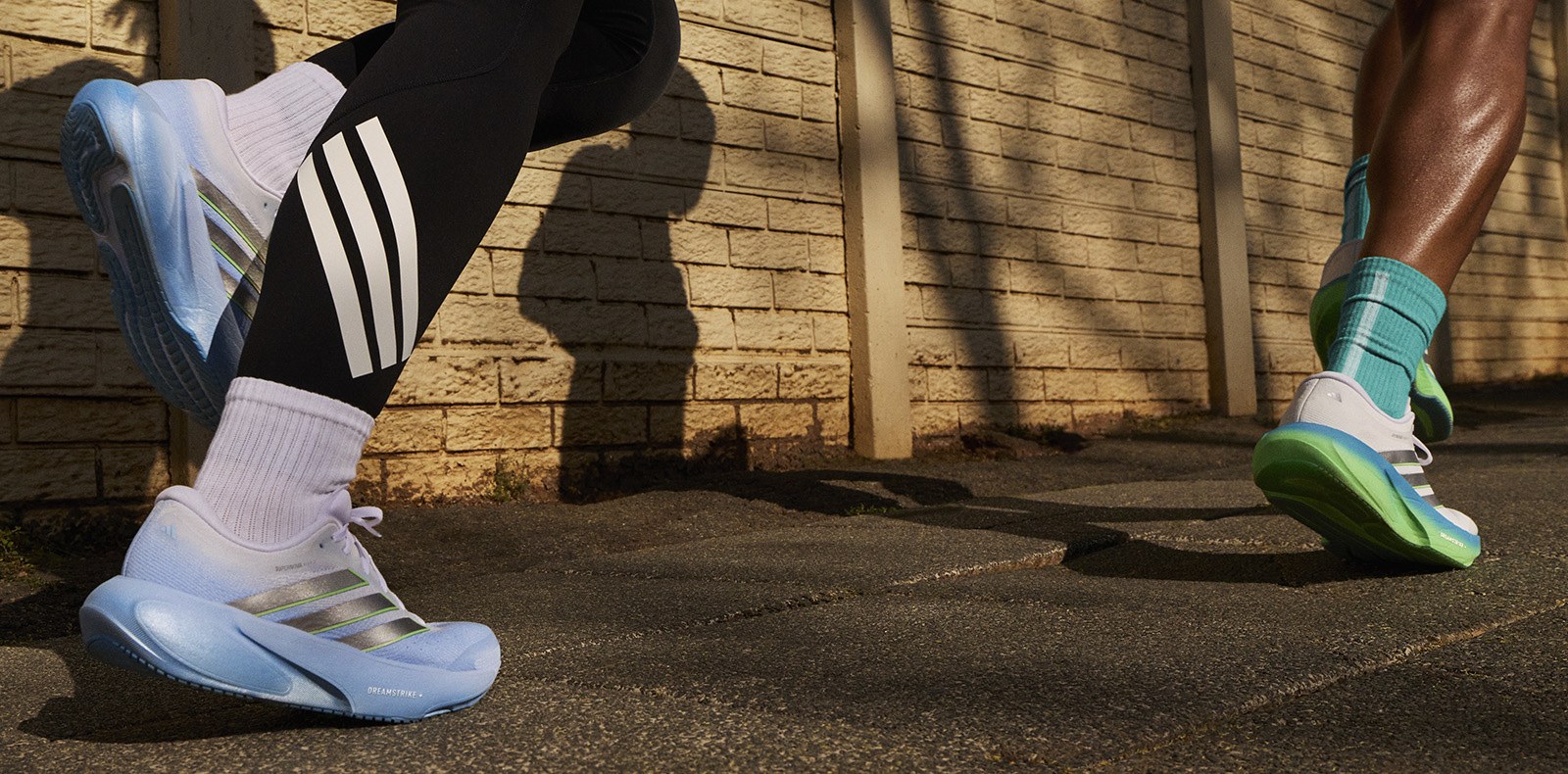 Colorful athletic shoes stand on a concrete surface, with a wooden structure and shadows in the background.