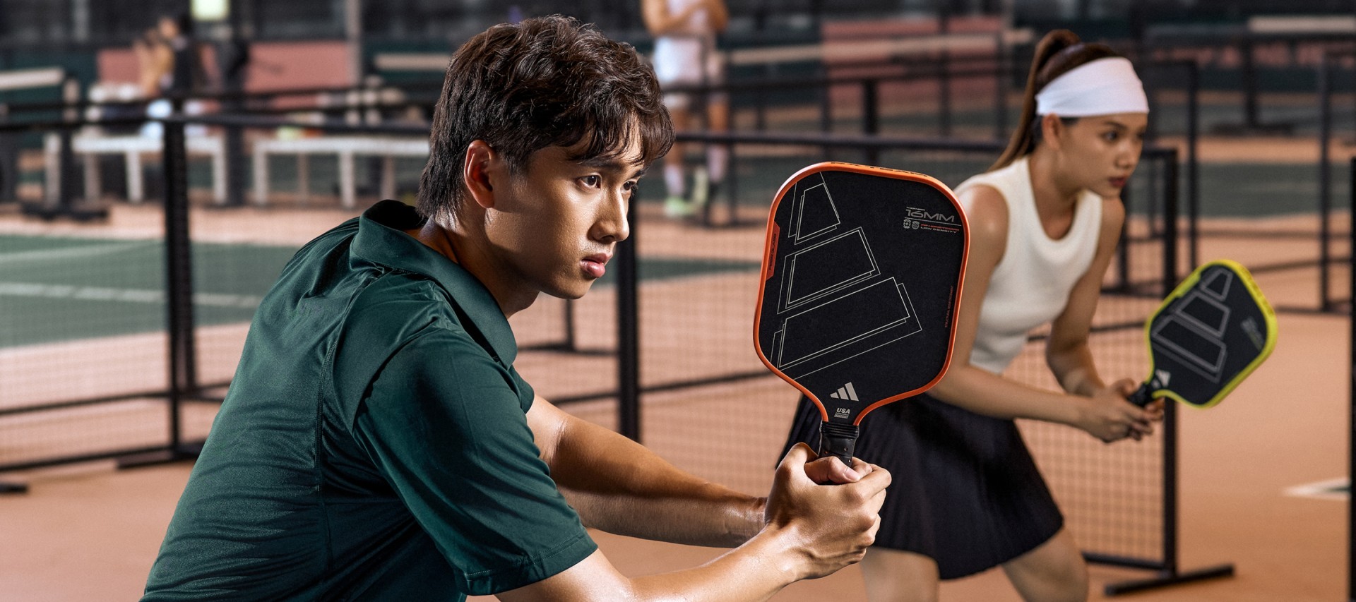 Two individuals, one wearing a green jacket and the other a white shirt, are engaged in a tennis match on a tennis court with a net visible in the background.