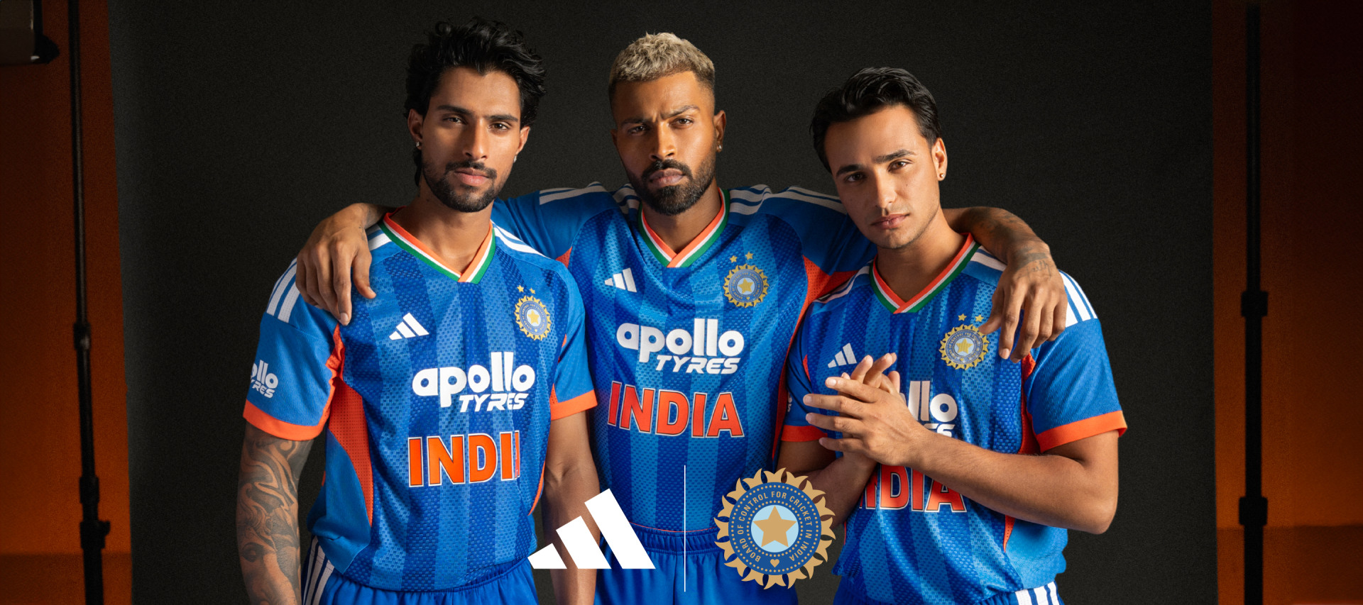 Three individuals wearing blue and orange cricket jerseys with the text "INDIA" and "apollo tyres" prominently displayed, standing against a dark background.