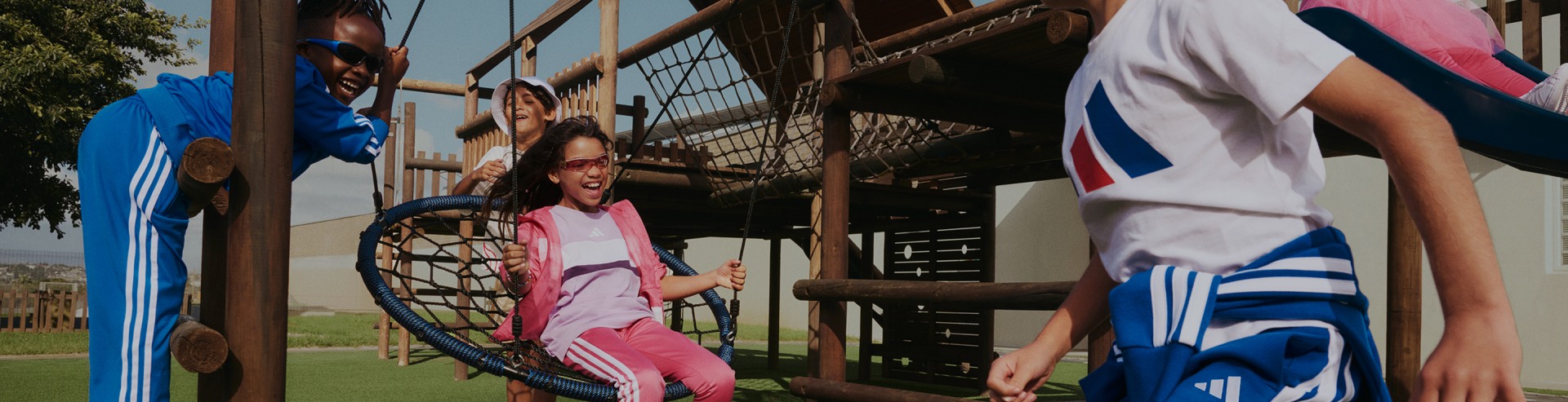 Two young girls, one wearing a pink outfit and the other in a blue outfit, are playing on a wooden and rope playground structure in an outdoor setting.