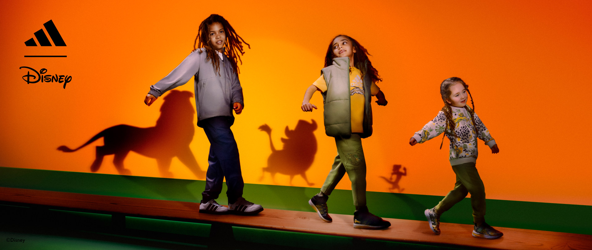Three children walking across a bench followed by Lion King shadows, wearing the adidas | Lion King collection.