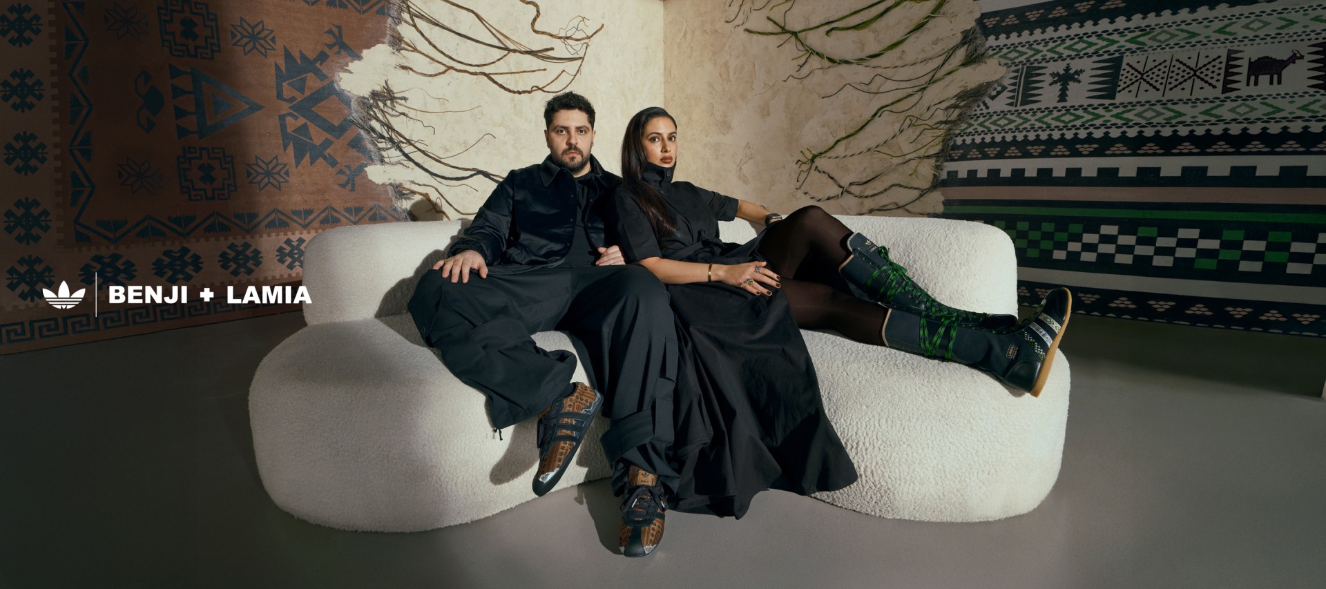 Two individuals, a man and a woman, are seated on a couch against a backdrop of intricate, patterned walls and decorations.