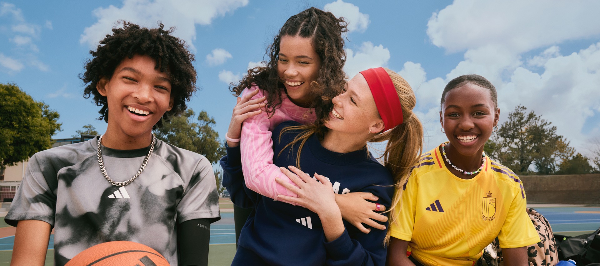 A group of four young, smiling individuals, dressed in casual attire, stand together in an outdoor setting with a cloudy sky and trees in the background.