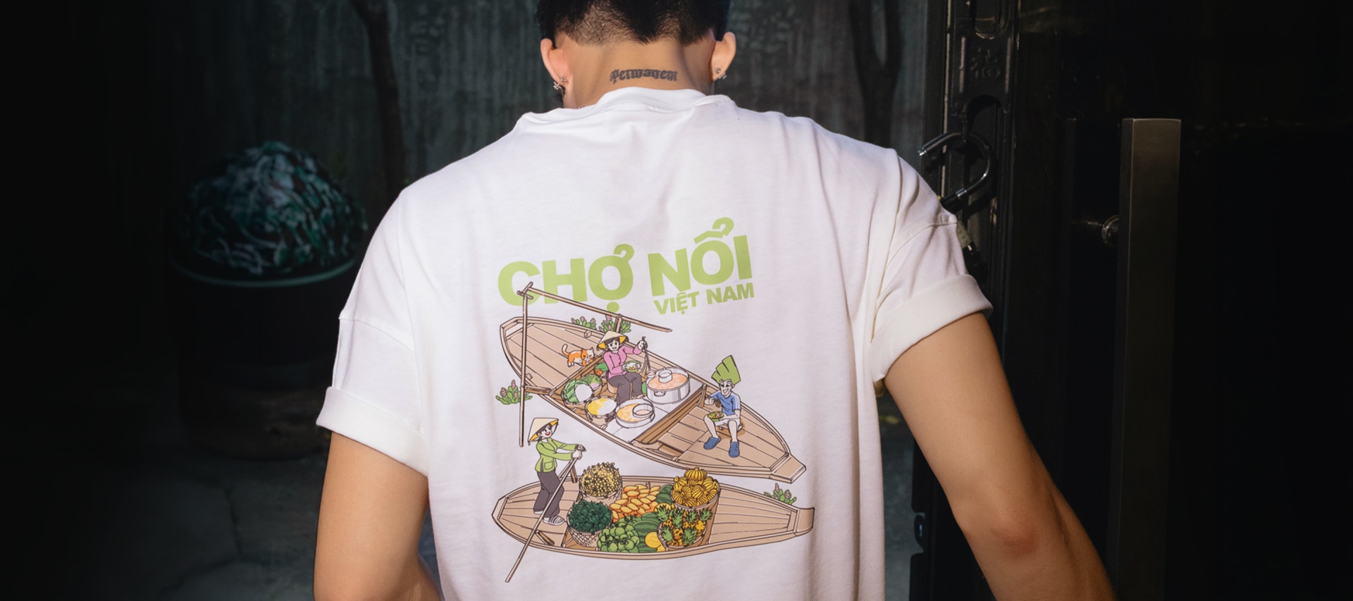 A person wearing a white t-shirt with a colorful graphic design depicting a boat filled with various characters and the text "Cho noi" in the background.