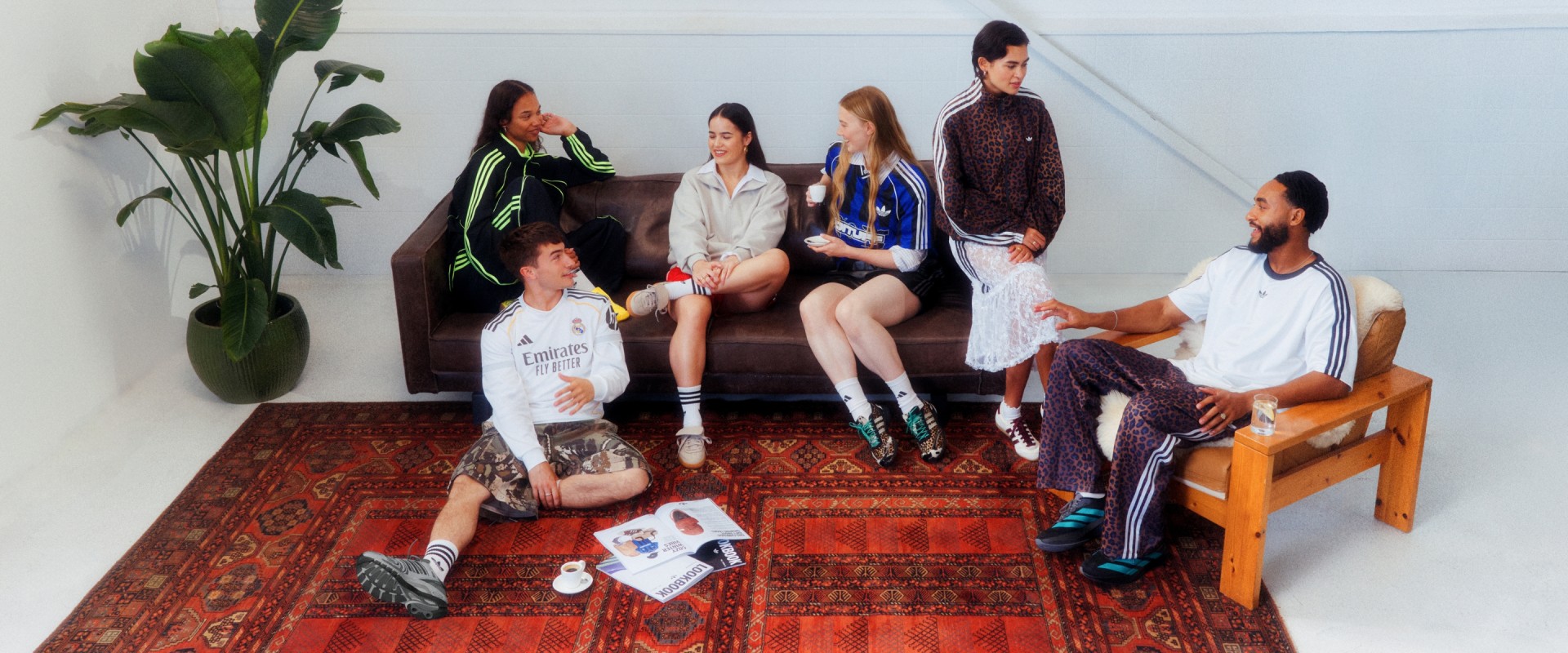 En grupp människor, en del sittande och en del stående, samlas på en färgstark mönstrad matta i vad som verkar vara en inomhusmiljö med krukväxter i bakgrunden.