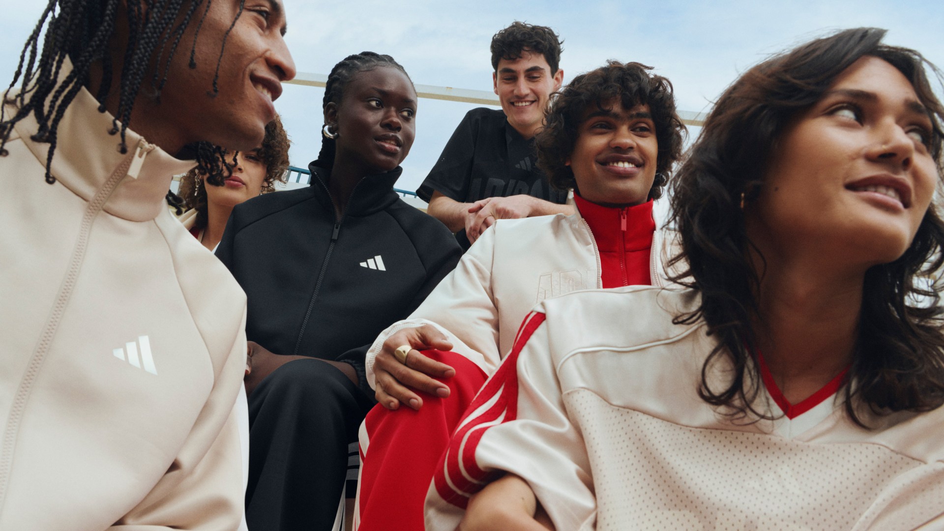 A group of young people, some wearing casual clothing and others in athletic attire, are gathered together in an outdoor setting with a clear sky in the background.