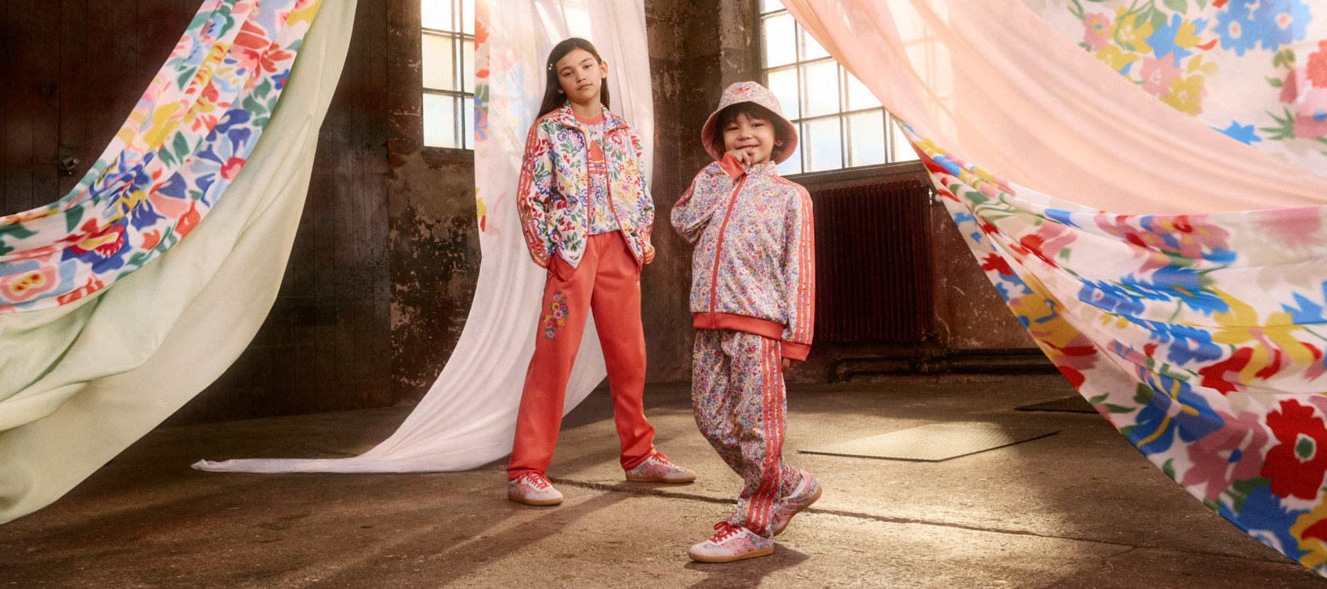 A girl and boy model the adidas Originals x Liberty London collection in an industrial space decorated with flowing, patterned fabric.