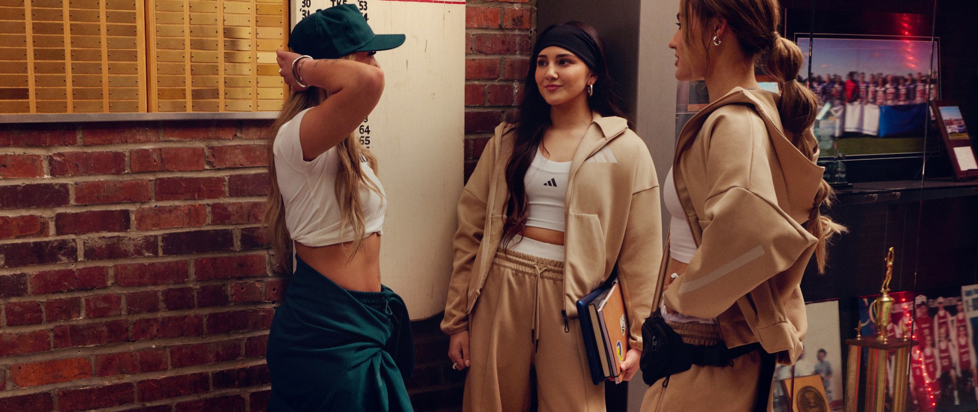 Three young women stand talking in the hall of a sports college with leader boards and medals in the background, wearing apparel from the new ADIDAS Z.N.E. range. 