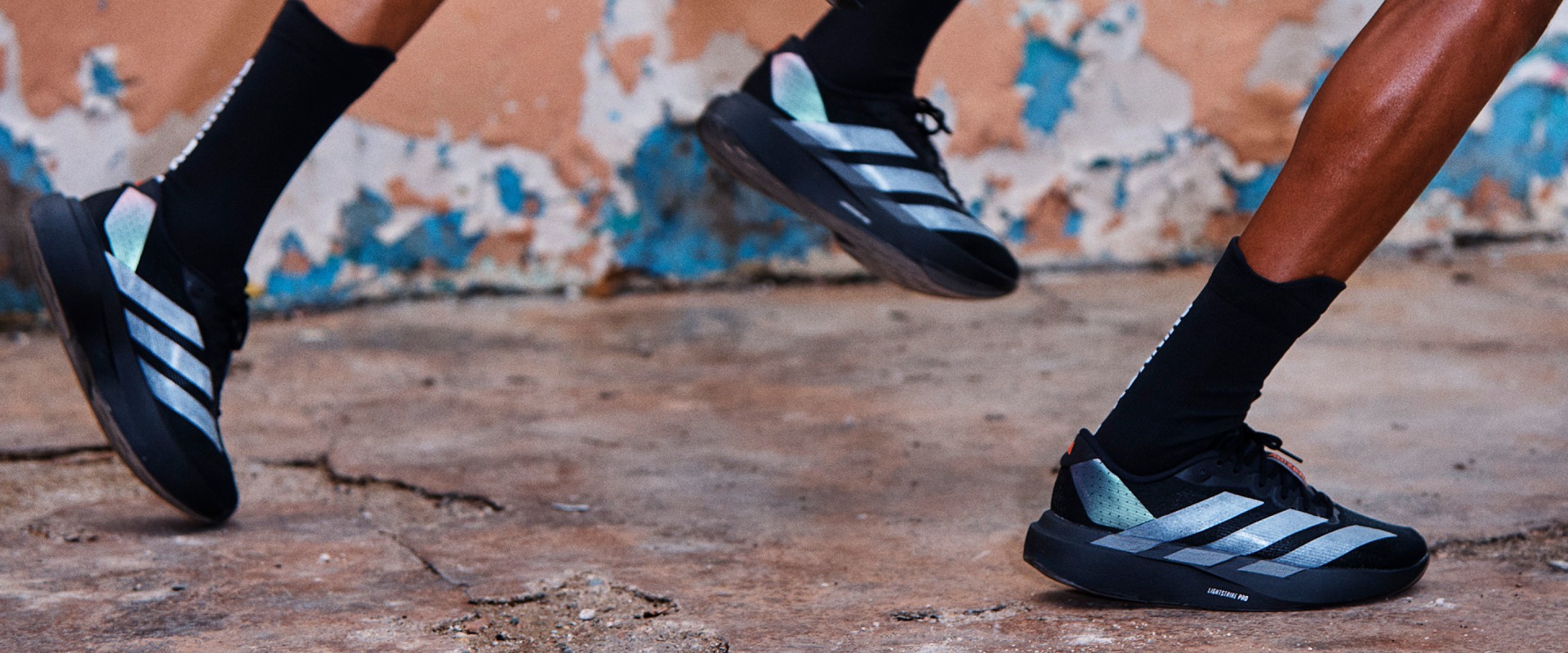 The image shows a pair of athletic shoes with a colorful, patterned design, standing on a rocky, uneven surface in the foreground, with a blurred background suggesting an outdoor setting.