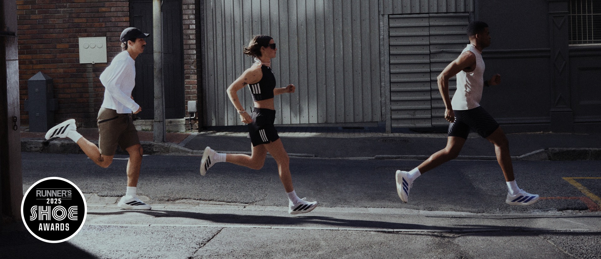 Three people in athletic wear are running on a sidewalk in front of a brick building with a metal door.