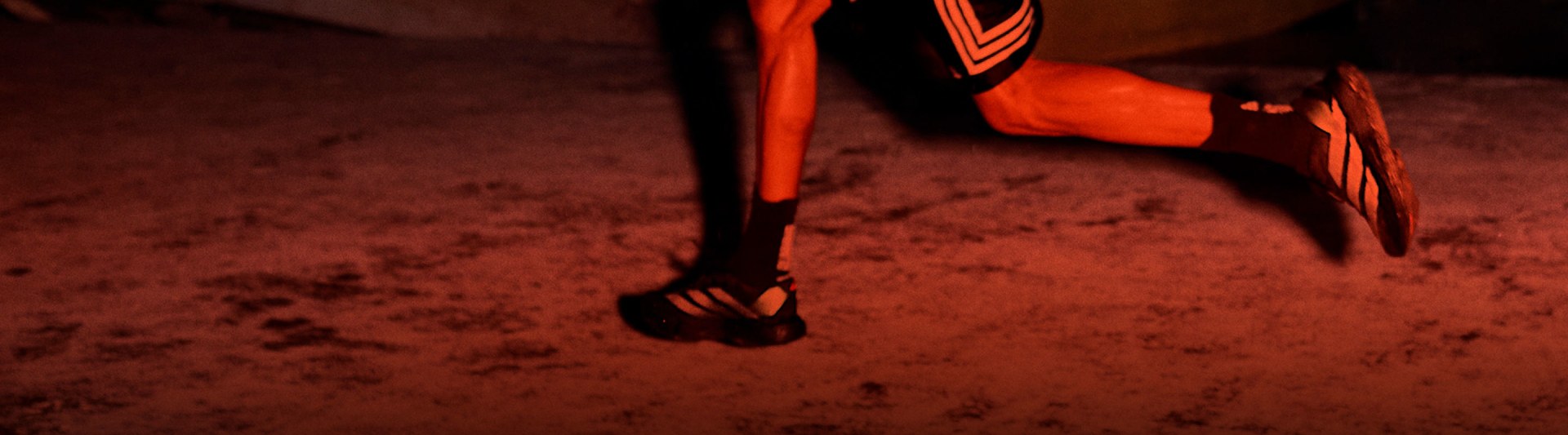 The image shows a close-up view of a person's lower legs and feet wearing bright orange athletic shoes or cleats on a reddish-brown surface, likely a sports field or court.