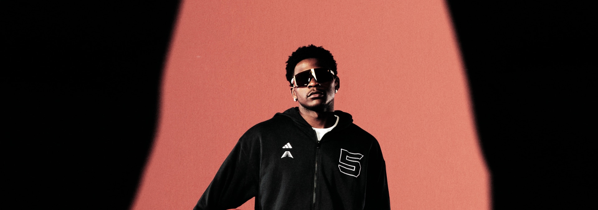 Anthony Edwards wears a black adidas tracksuit and sunglasses. He holds a basketball against a peach background.