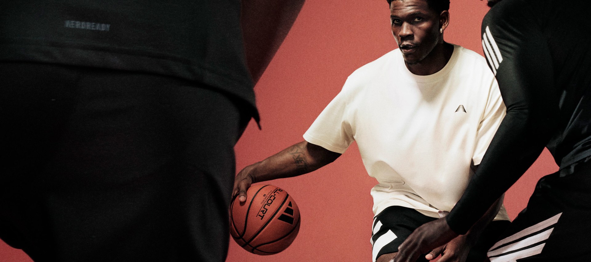 Anthony Edwards dribbles a basketball on an indoor court, wearing a white adidas t-shirt and black adidas shorts. Two defenders in black adidas outfits face him.