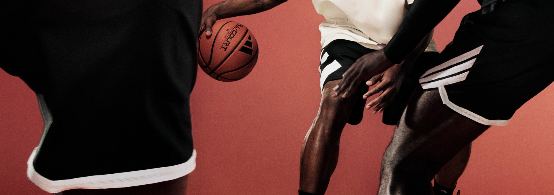 Anthony Edwards dribbles a basketball on an indoor court, wearing a white adidas t-shirt and black adidas shorts. Two defenders in black adidas outfits face him.