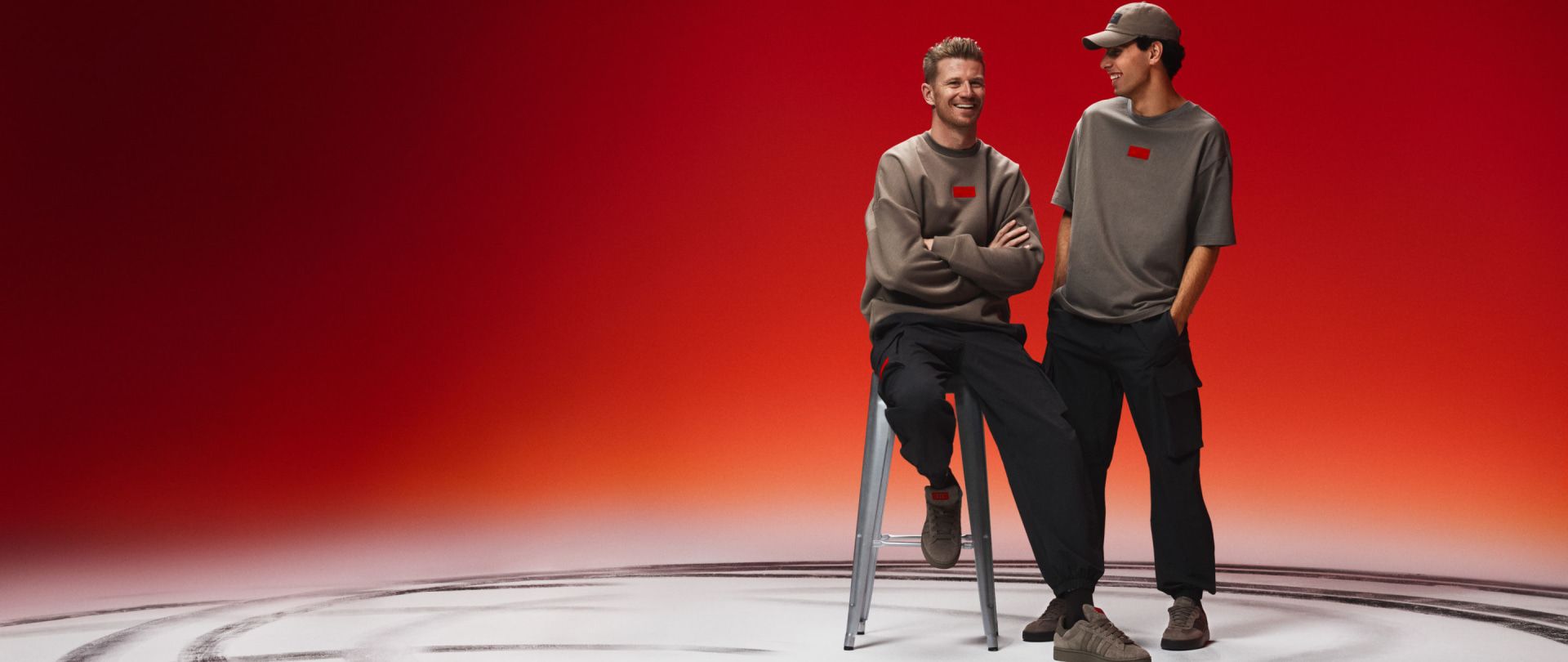 Two individuals wearing gray shirts and black pants stand on a platform against a vibrant red background.