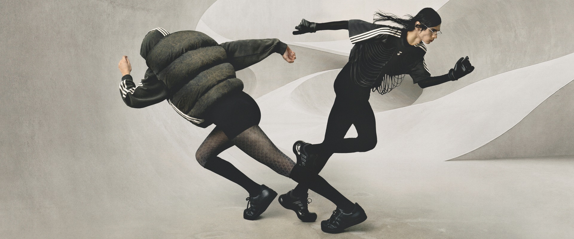 Two models running in opposite directions wearing adidas Originals x Avavav collection, including black puffer vest, sleeveless hoodie, and black Moonrubber trainers, set against a sculptural abstract concrete background.
