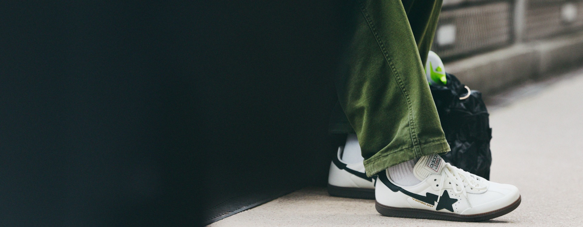 A close-up view of a person's lower body, featuring a pair of white trainers with a star design and a green pair of trousers or trousers in the foreground, with a dark background.