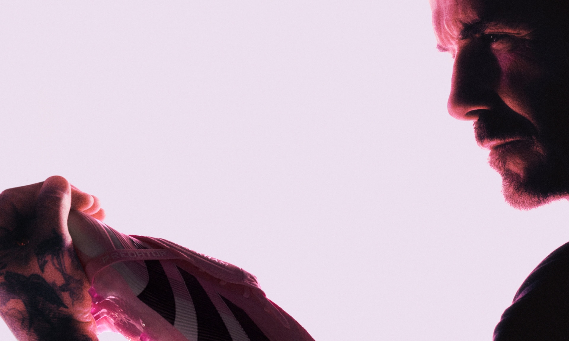 Beckham, dressed in black, gazes at a pink adidas Predator Elite FT FG cleat balanced in his hand against a neon pink backdrop.