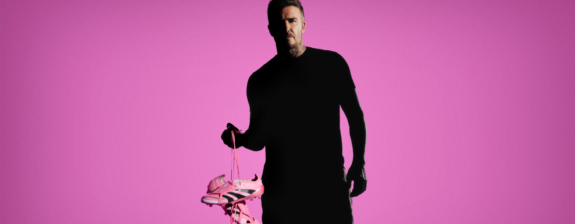 David Beckham stands in all-black, holding a pair of adidas Predator Elite FT FG cleats by the laces against a neon pink backdrop.