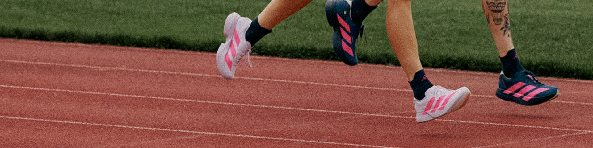 The image shows a close-up view of a person's legs and feet running on a red running track, with a grassy field in the background.