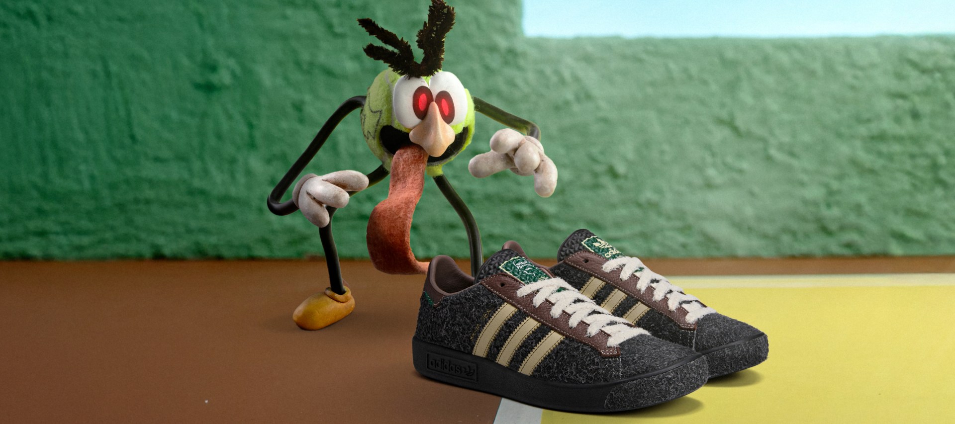 Brown adidas Originals x Brain Dead sneakers styled beside a quirky, tennis ball-shaped monster, set on wooden floors against a green wall.