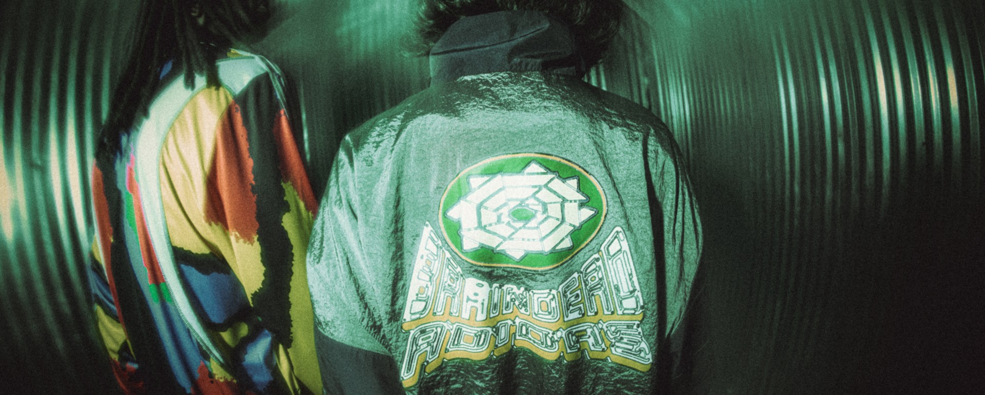 A hooded figure wearing a jacket with a colorful, geometric design stands in a dimly lit, green-tinted environment.