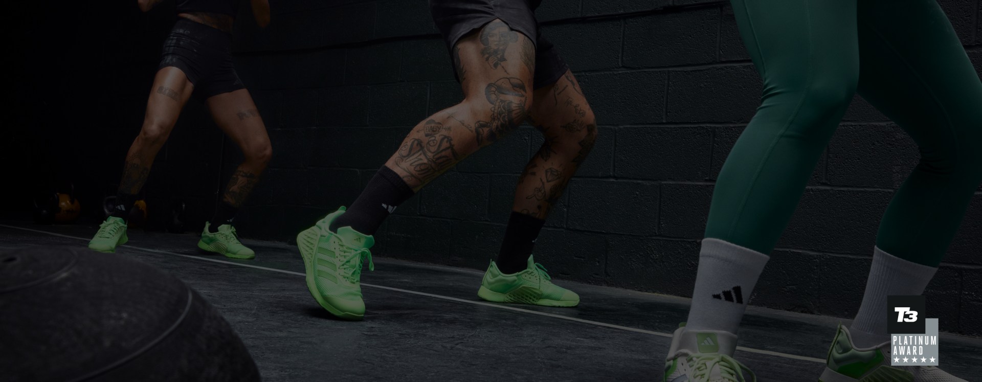 A man with dark hair lifting weights in black adidas workout clothes and green Dropset 3 shoes at the gym.