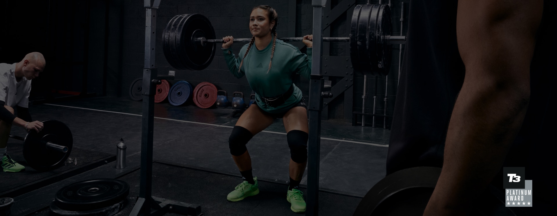 A man with dark hair lifting weights in black adidas workout clothes and green Dropset 3 shoes at the gym.