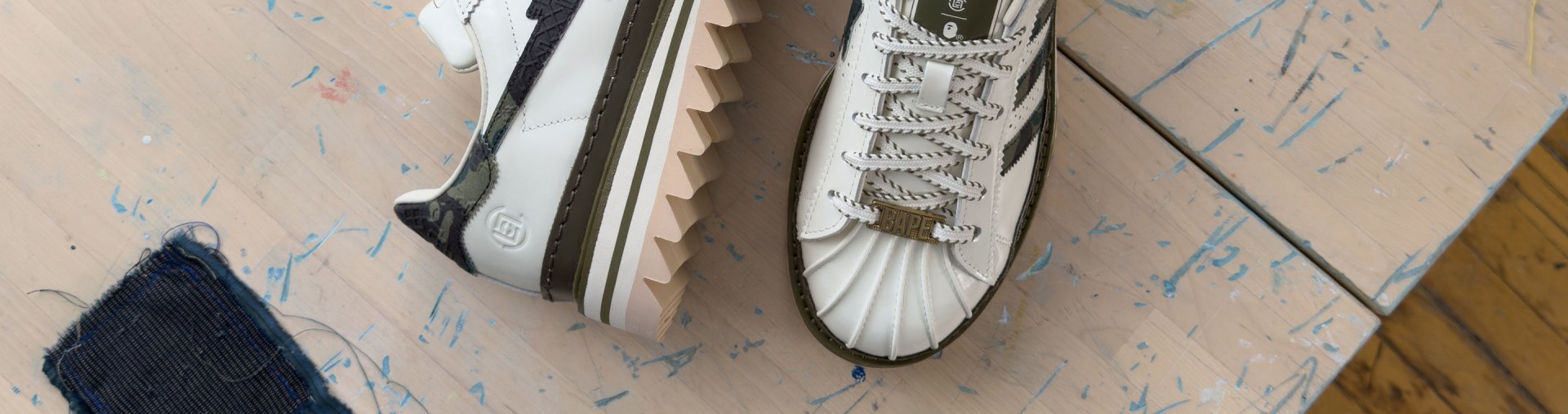 Um par de tênis adidas CLOT x BAPE Superstar em uma superfície leve de madeira com marcas de tinta azul. Um dos tênis exibe o logo da estrela preta e o outro exibe a etiqueta adidas e as Três Listras.