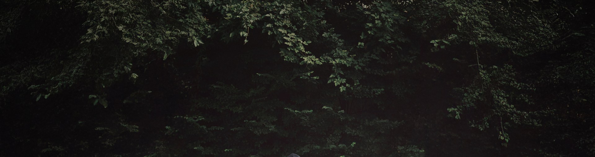 A person sits on a wooden bench outdoors, wearing adidas CLOT Pro Model Collection by Edison Chen consisting of a black adidas jacket with patches, light-coloured trousers, dark trainers, and a black cap.