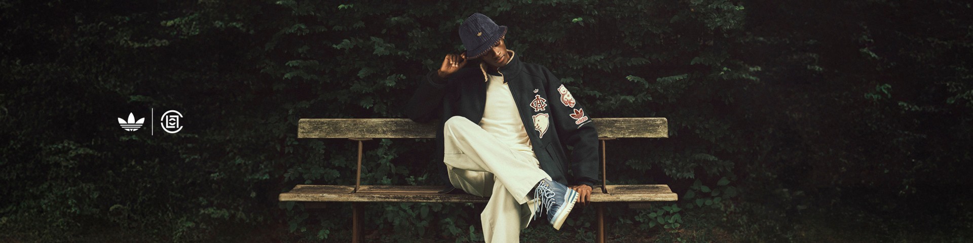 Un hombre sentado en un banco de madera al aire libre, usando la colección adidas CLOT Pro Model by Edison Chen que consiste en un gorro pescador azul marino, una chaqueta negra con parches bordados, pantalones crema y zapatillas adidas azules.