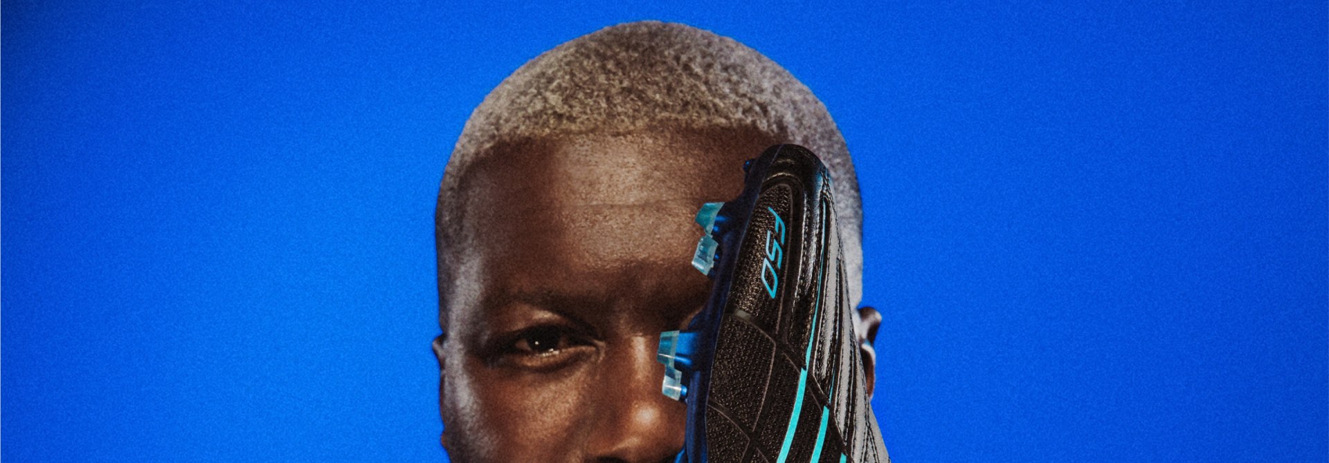 鮮やかなブルーを背景に、ジブリル・シセが顔の前で黒いジャケットを着てブーツを持つ姿をクローズアップしたポートレート。