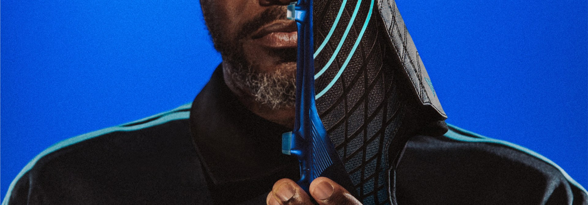 A close-up portrait of a person with a beard, wearing a dark hooded garment against a vibrant blue background.