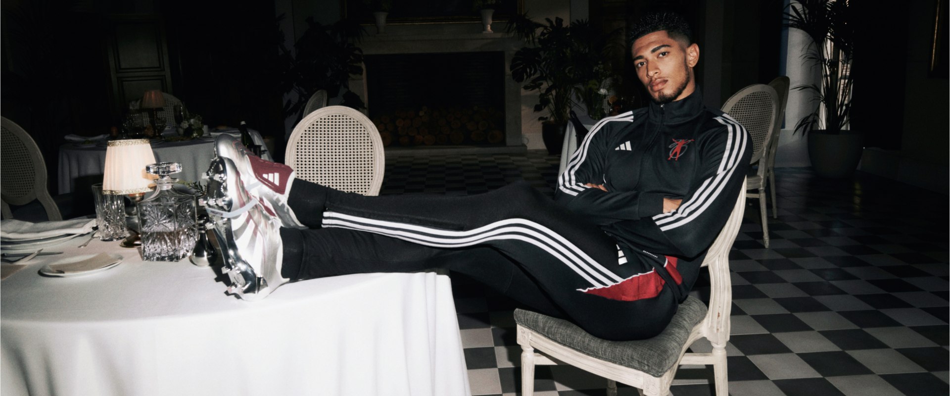 Jude Bellingham wearing a black and white athletic outfit is sitting on a chair in a room with a checkered floor and various decorative items in the background.