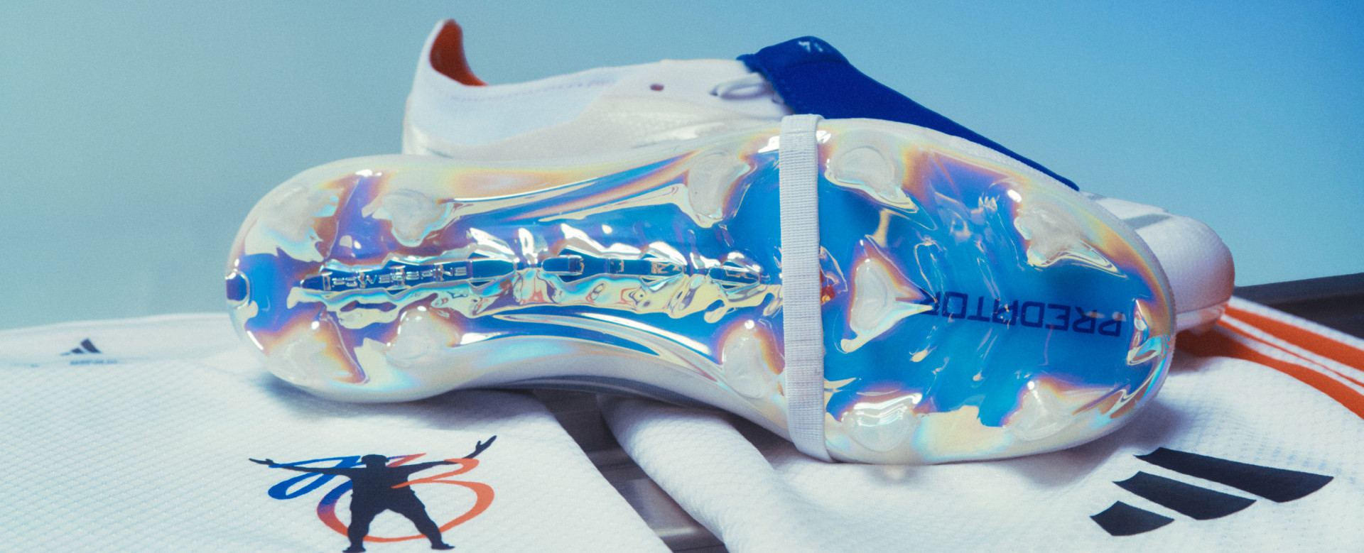 A pair of adidas Jude Bellingham Predator boots rest on a black bench, set against a cool blue gradient background.