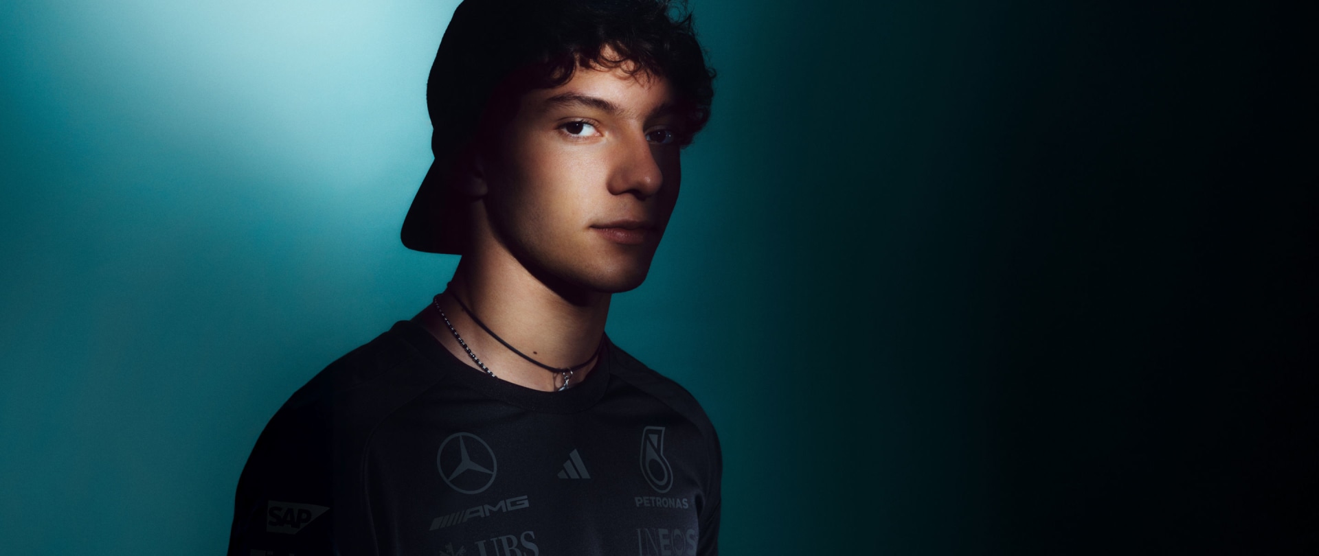 A young person with dark hair and a pensive expression, set against a teal-colored background.