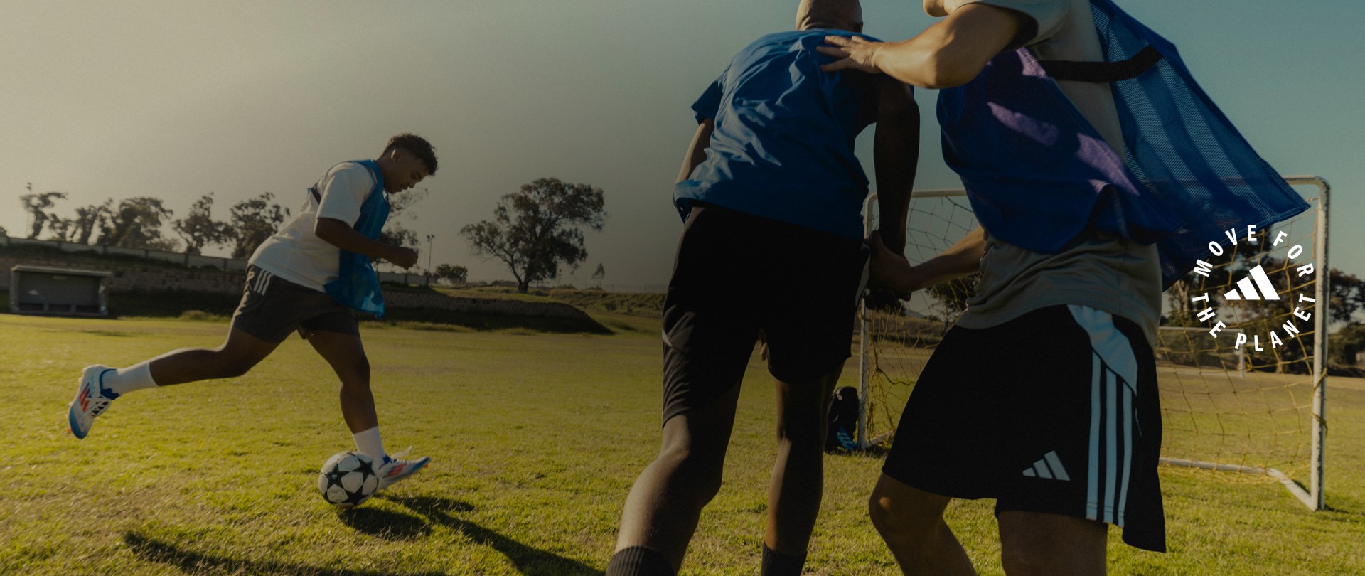 The image shows two people playing soccer on a grassy field, with trees and a building visible in the background.