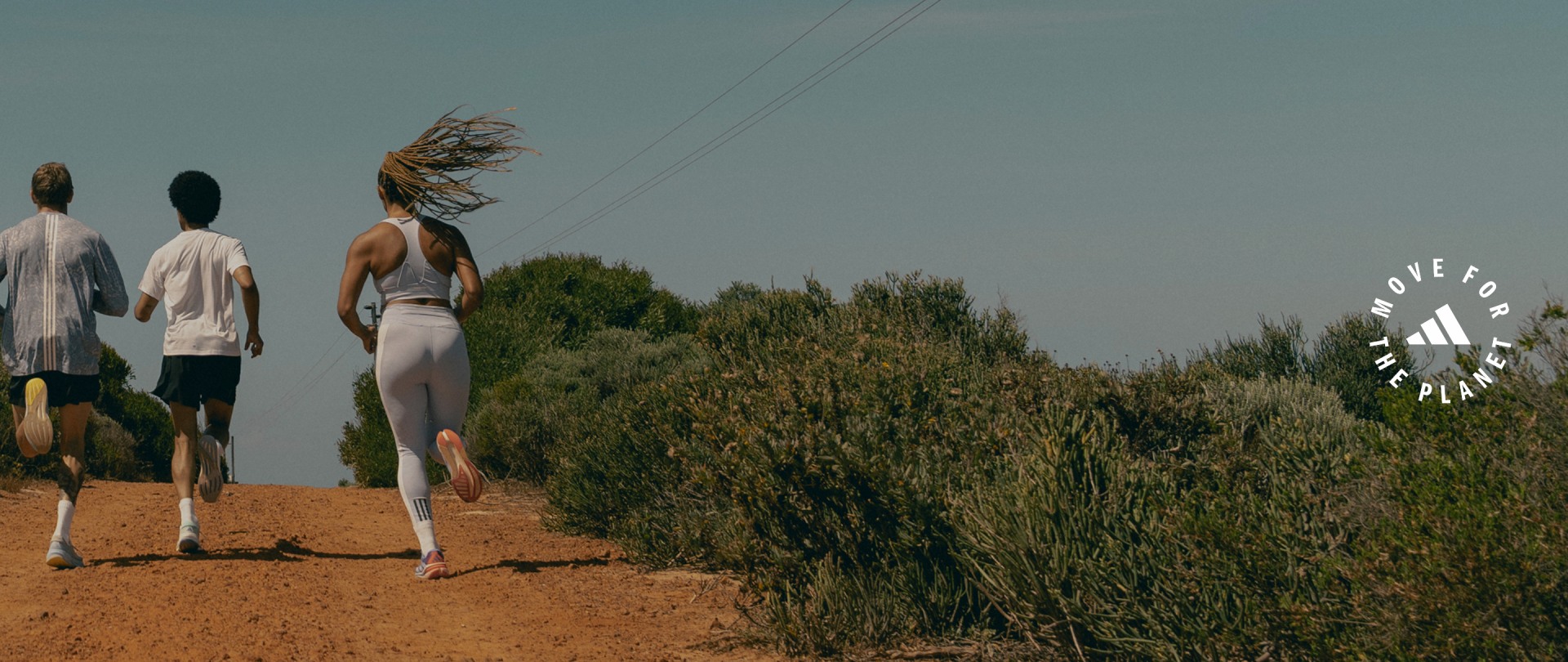 Two people, one with long hair, stand on a dirt path surrounded by lush vegetation against a clear sky.