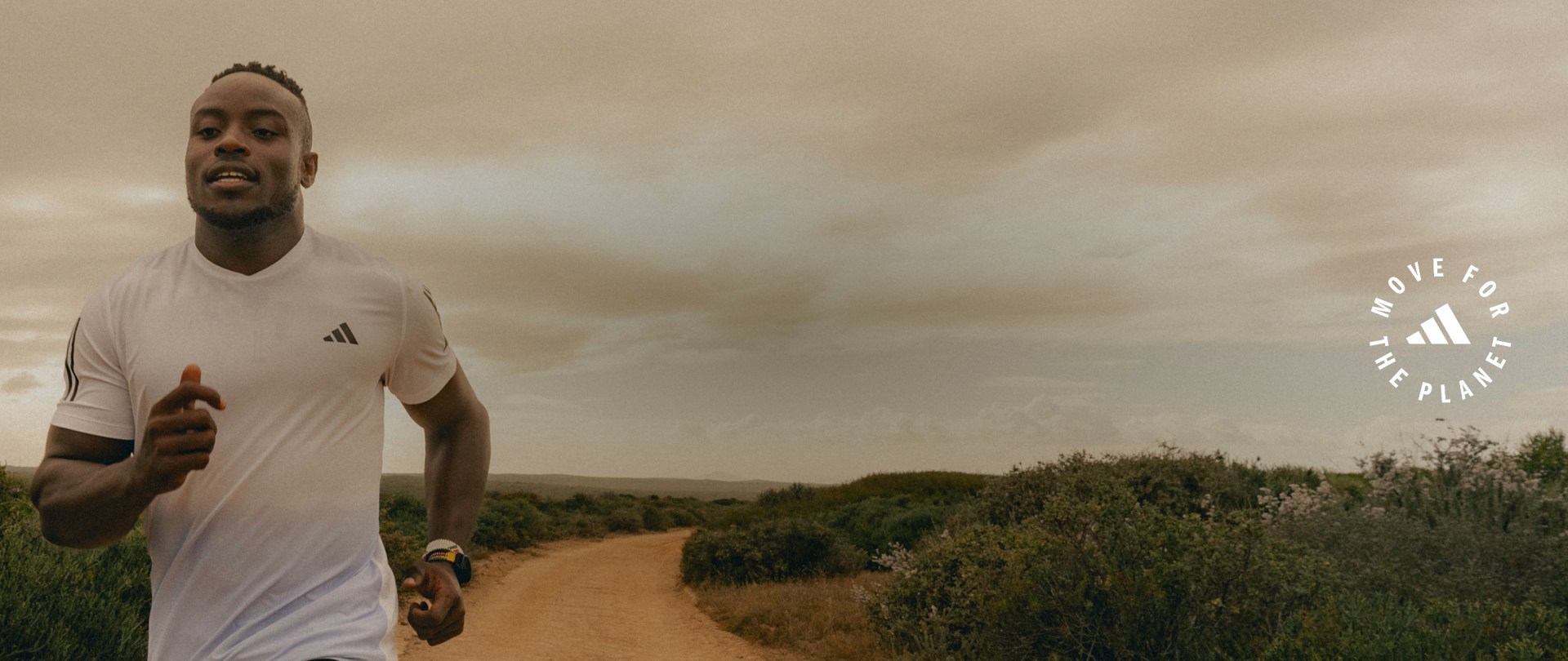 A person wearing a white athletic shirt stands on a dirt path surrounded by lush greenery and a cloudy sky in the background.