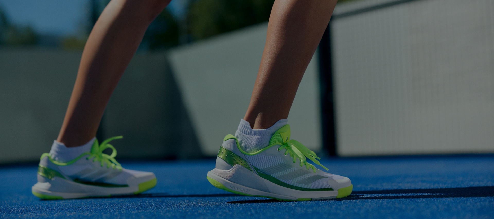 The image shows a pair of athletic shoes with green and white accents, standing on a blue padel court.